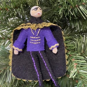 May include: A handmade Christmas ornament in the shape of a figure with a painted face, eye patch, and mustache. The figure wears a purple jacket, black pants with purple accents, and a black cape with gold trim. The ornament hangs from a gold string.