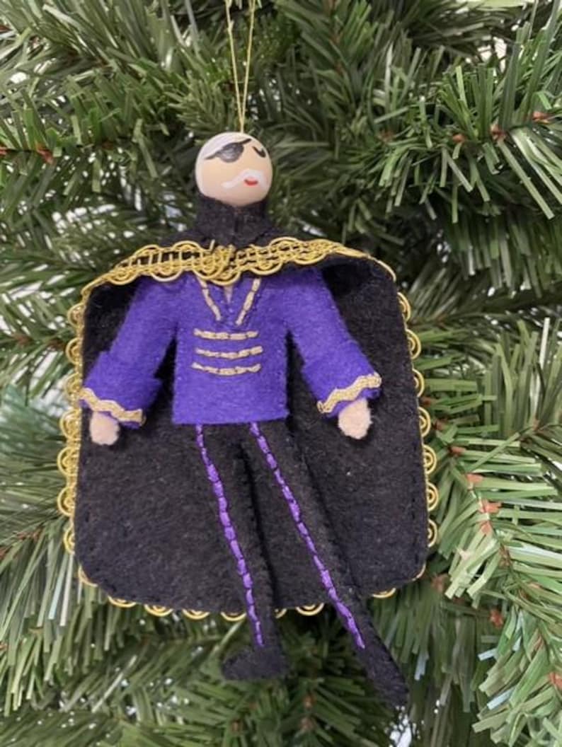 May include: A handmade Christmas ornament in the shape of a figure with a painted face, eye patch, and mustache. The figure wears a purple jacket, black pants with purple accents, and a black cape with gold trim. The ornament hangs from a gold string.