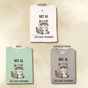 May include: Three t-shirts in ivory, bay, and sandstone colors. Each shirt features a graphic of a cartoon raccoon with the text "NOT AI." above and "JUST BUILT DIFFERENT" below the raccoon. The shirts have a crew neck and short sleeves.