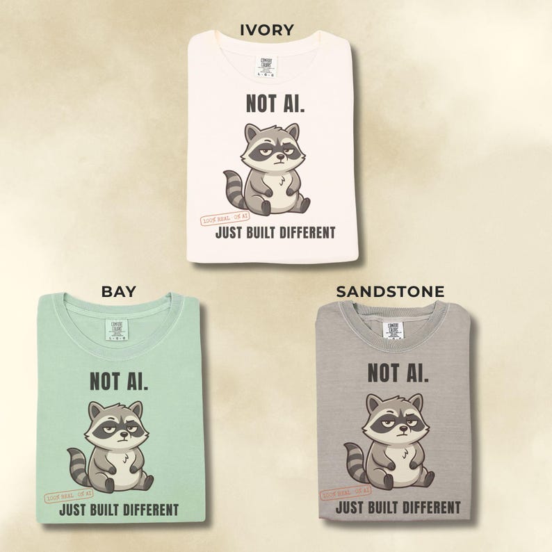 May include: Three t-shirts in ivory, bay, and sandstone colors. Each shirt features a graphic of a cartoon raccoon with the text "NOT AI." above and "JUST BUILT DIFFERENT" below the raccoon. The shirts have a crew neck and short sleeves.