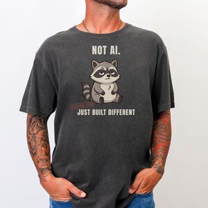 May include: A charcoal gray t-shirt featuring a graphic of a sitting raccoon with a skeptical expression. Above the raccoon is the text NOT AI. Below the raccoon is a small red stamp and the text JUST BUILT DIFFERENT in white capital letters.