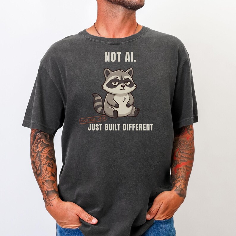 May include: A charcoal gray t-shirt featuring a graphic of a sitting raccoon with a skeptical expression. Above the raccoon is the text NOT AI. Below the raccoon is a small red stamp and the text JUST BUILT DIFFERENT in white capital letters.
