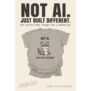 May include: A light gray t-shirt featuring a cartoon raccoon graphic with the text "NOT AI." above and "JUST BUILT DIFFERENT" below. The shirt includes a small red stamp graphic that reads "100% REAL - 0% AI" near the raccoon illustration.