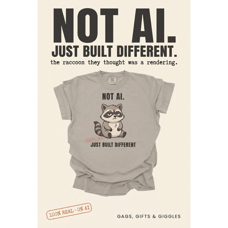 May include: A light gray t-shirt featuring a cartoon raccoon graphic with the text "NOT AI." above and "JUST BUILT DIFFERENT" below. The shirt includes a small red stamp graphic that reads "100% REAL - 0% AI" near the raccoon illustration.