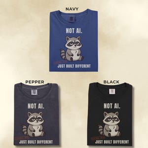 May include: Three t-shirts in navy, pepper, and black colors. Each shirt features a graphic of a cartoon raccoon with the text "NOT AI." above and "JUST BUILT DIFFERENT" below the raccoon, along with a small "100% REAL" stamp.