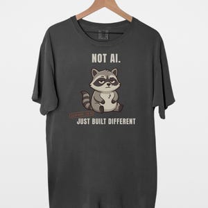 May include: A charcoal gray short-sleeve t-shirt featuring a cartoon raccoon illustration. The text above the raccoon reads NOT AI. and the text below reads JUST BUILT DIFFERENT. The raccoon has a gray and white face with a striped tail.