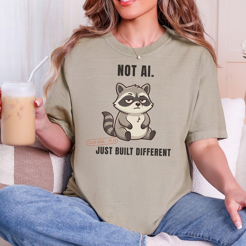 May include: A light olive green t-shirt featuring a cartoon raccoon graphic in the center. Above the raccoon, the text reads NOT AI. Below the raccoon, the text reads JUST BUILT DIFFERENT. The shirt has a crew neckline and short sleeves.