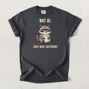 May include: A charcoal gray t-shirt featuring a cartoon raccoon graphic in the center. Above the raccoon is the text NOT AI. Below the raccoon is the text JUST BUILT DIFFERENT. The shirt has short sleeves and a crew neckline with a visible brand label.