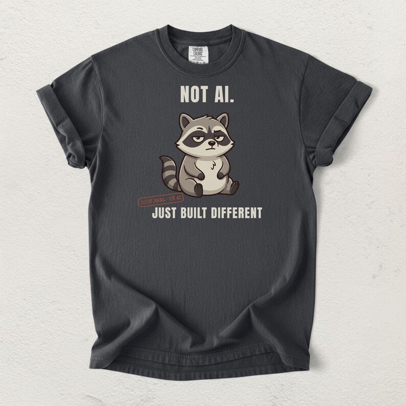 May include: A charcoal gray t-shirt featuring a cartoon raccoon graphic in the center. Above the raccoon is the text NOT AI. Below the raccoon is the text JUST BUILT DIFFERENT. The shirt has short sleeves and a crew neckline with a visible brand label.