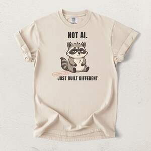 May include: A beige short-sleeve t-shirt featuring a graphic of a grey and white raccoon with a skeptical expression. The text reads NOT AI. above the raccoon and JUST BUILT DIFFERENT below it, with a small stamp stating 100% REAL 0% AI.