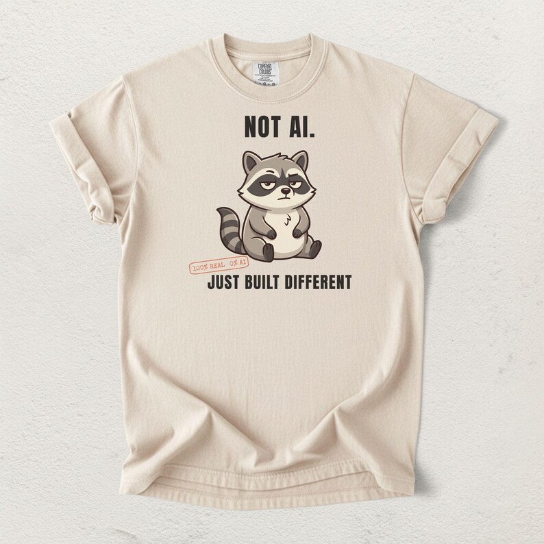 May include: A beige short-sleeve t-shirt featuring a graphic of a grey and white raccoon with a skeptical expression. The text reads NOT AI. above the raccoon and JUST BUILT DIFFERENT below it, with a small stamp stating 100% REAL 0% AI.