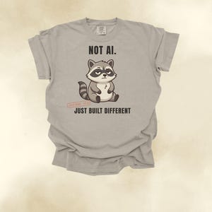 May include: A light gray t-shirt featuring a cartoon raccoon illustration in the center. Above the raccoon is the text "NOT AI." and below it is the text "JUST BUILT DIFFERENT." A small red stamp graphic near the raccoon reads "100% REAL. 0% AI."