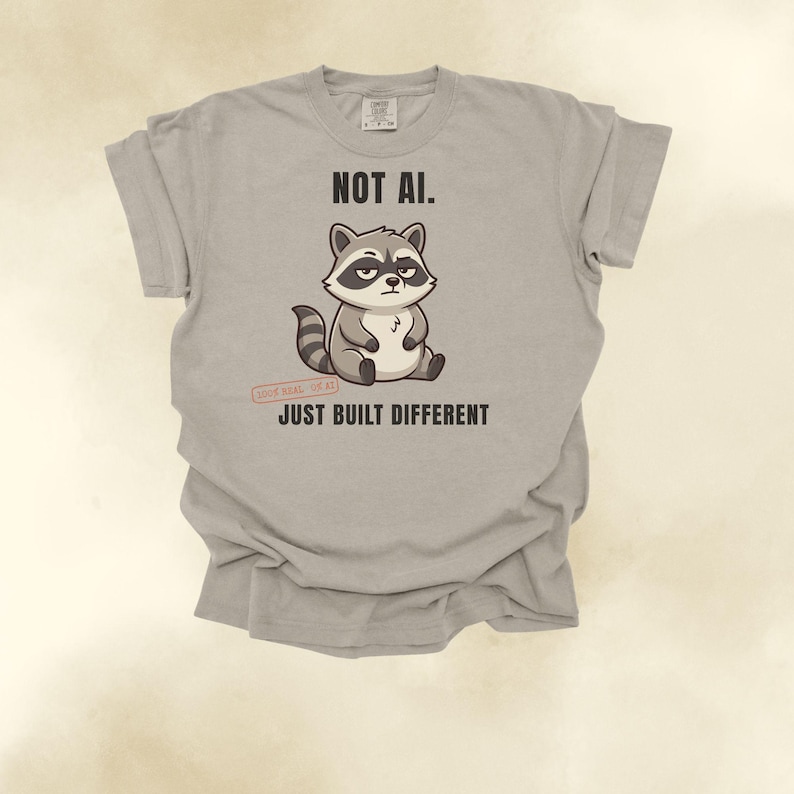 May include: A light gray t-shirt featuring a cartoon raccoon illustration in the center. Above the raccoon is the text "NOT AI." and below it is the text "JUST BUILT DIFFERENT." A small red stamp graphic near the raccoon reads "100% REAL. 0% AI."