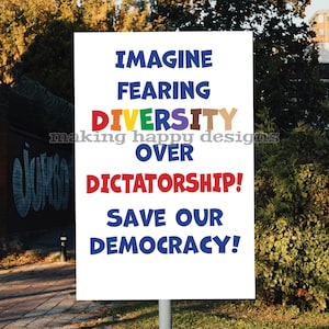Op de afbeelding: Een wit bord met de tekst "IMAGINE FEARING DIVERSITY OVER DICTATORSHIP! SAVE OUR DEMOCRACY!" in blauw, rood en regenboogkleuren. Het bord wordt vastgehouden door een metalen paal, met een vage achtergrond van bomen en een bakstenen muur.