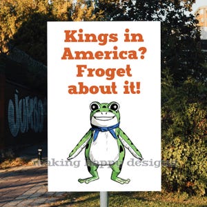 May include: A white sign with the text "Kings in America? Froget about it!" in orange. The sign features a cartoon frog with a blue scarf. The frog is green with black spots and a white belly. The sign is outdoors.