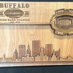 May include: Wooden wall art featuring a laser-cut design of Buffalo, New York. The artwork includes the city skyline, Highmark Stadium, and the text "BUFFALO" and "HIGHMARK STADIUM EST. 1872". The piece is made of light-colored wood.