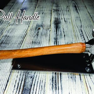 Pode incluir: Uma puxador com cabo de madeira e cabeça de martelo em aço inoxidável. A pega é fixada a uma placa de metal preta. O texto "Pull Handle" está escrito em uma fonte cursiva.
