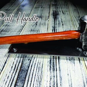 May include: A metal pull handle with a wooden handle and a hammer head. The handle is attached to a rectangular metal base with screws. The text "Pull Handle" is written in a script font.