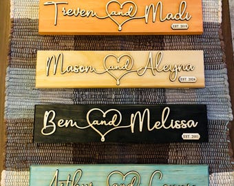 May include: Four wooden signs with the names of couples and their wedding dates. The signs are painted in different colors: orange, light brown, black, and light blue. The names are written in cursive with a heart symbol between them. The signs are perfect for a wedding or anniversary gift.