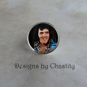 May include: A round, silver-toned pin featuring a smiling image of Elvis Presley in a blue jacket and patterned scarf. The pin has the text "Designs by Chastity" in a cursive font.