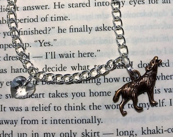 May include: Silver-toned chain bracelet with a heart-shaped crystal charm and a bronze-colored howling wolf pendant. The bracelet is displayed on an open book.