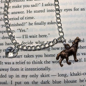 May include: Silver-toned chain bracelet with a heart-shaped crystal charm and a bronze-colored howling wolf pendant. The bracelet is displayed on an open book.