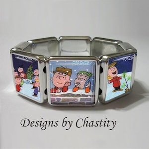 以下が含まれることがあります： 銀色のストレッチブレスレット。正方形のパネルには、冬のシーンで漫画のキャラクターが描かれています。パネルにはチャーリー・ブラウンと仲間たちが描かれており、「FOREVER」の文字と「Designs by Chastity」の文字があります。