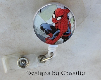 May include: A round, white retractable badge reel featuring a comic book image of Spider-Man in a red and blue suit. The reel has a clear plastic strap and a metal clip. Text on the reel reads "Designs by Chastity."