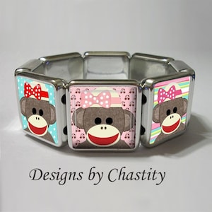 May include: A silver-toned stretch bracelet featuring square charms with sock monkey designs. Each charm showcases a brown monkey face with a red mouth, black eyes, and a bow. The background colors vary, including blue, pink, and striped patterns. Text: Designs by Chastity.