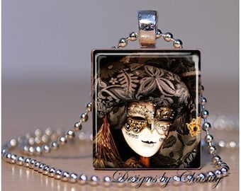 May include: A square pendant necklace featuring a detailed image of a Venetian masquerade mask. The mask is white and gold, set against a dark, patterned background. The necklace has a silver-colored chain and a metal bail.