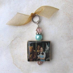 May include: A rectangular pendant featuring a scene from The Wizard of Oz, with a silver-tone ring, pink and blue beads, and a gold ribbon. The image shows Dorothy, the Scarecrow, the Tin Man, and the Cowardly Lion.