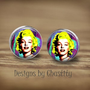 Puede incluir: Un par de pendientes redondos con una imagen de estilo pop art de Marilyn Monroe con cabello amarillo, sobre un fondo geométrico colorido. Los pendientes se muestran sobre una superficie de madera. El texto "Designs by Chastity" es visible.