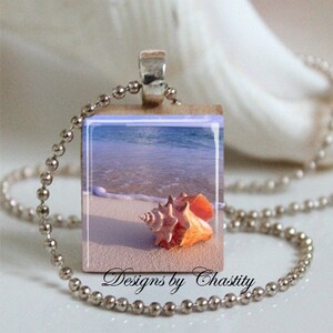 以下が含まれることがあります： ビーチの風景が描かれたペンダントが付いたシルバーカラーのネックレス。ペンダントには、砂浜に打ち上げられた貝殻と海の波が描かれています。「Designs by Chastity」の文字が見えます。