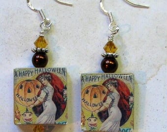 Pode incluir: Um par de brincos com tema de Halloween. Cada brinco apresenta um pingente quadrado com uma ilustração vintage de uma mulher segurando uma abóbora, com o texto "A Happy Halloween". Os brincos têm ganchos prateados, contas marrons e detalhes em cristal laranja.