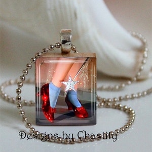 Puede incluir: Un collar con colgante cuadrado con una imagen de zapatos rojos, calcetines azules y una varita en forma de estrella. El colgante está en una cadena de bolas plateada. El texto "Designs by Chastity" está en la parte inferior.