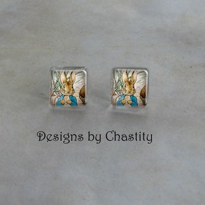 May include: Two square stud earrings featuring a watercolor illustration of Peter Rabbit in a blue jacket. The earrings have a clear, glossy finish. The text "Designs by Chastity" is below the earrings.
