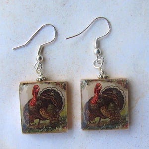 May include: A pair of square earrings featuring a vintage illustration of a turkey. The earrings have silver-colored hooks and a decorative border around the turkey image. The turkey is depicted with red, brown, and black feathers.