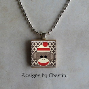 Könnte beinhalten: Eine quadratische Anhänger-Halskette mit einem Sockenaffen-Design. Der Affe hat einen rot-weiß gestreiften Hut, einen roten Mund und braunes Fell. Der Hintergrund ist ein braun-weißes Rautenmuster. Die Halskette ist silberfarben.