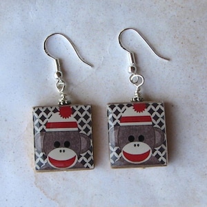 May include: A pair of square earrings featuring a sock monkey design. The earrings have a silver hook and a small silver bead. The sock monkey has a red and white hat and a red smile. The background is a black and white diamond pattern.