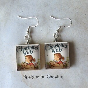 May include: Square earrings featuring the cover art of "Charlotte's Web." The image shows a young girl with red hair and a pig, with the title of the book above. The earrings have silver-colored fish hook ear wires.