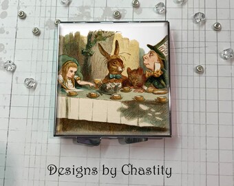 May include: Square compact mirror featuring a colorful illustration of the Mad Hatter's tea party from Alice in Wonderland. The mirror has a silver metal frame and the text "Designs by Chastity" at the bottom.