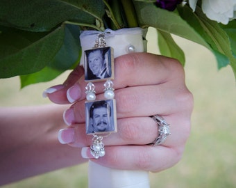May include: A bridal bouquet charm featuring two small square pendants with black and white photos of men. The pendants are connected by a silver chain with pearls and crystals. The charm is held in a hand with a diamond ring.