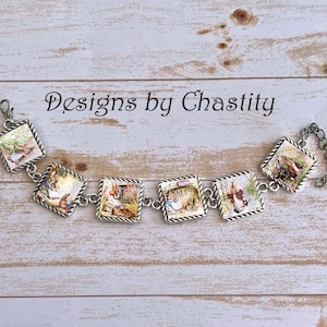 May include: Silver-toned bracelet featuring square charms with illustrations of rabbits in a garden setting. Each charm is framed with a twisted rope design. The bracelet is connected by small chain links. The text "Designs by Chastity" is above the bracelet.