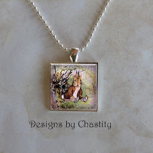 May include: A silver-toned necklace featuring a square pendant with a vintage illustration of a brown rabbit in a jacket. The pendant is attached to a ball chain. The text "Designs by Chastity" is below the pendant.