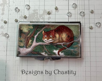 May include: A rectangular metal case featuring a colorful illustration of the Cheshire Cat from Alice in Wonderland, perched on a tree branch. The cat has a wide grin and is set against a forest backdrop. The text "Designs by Chastity" is at the bottom.