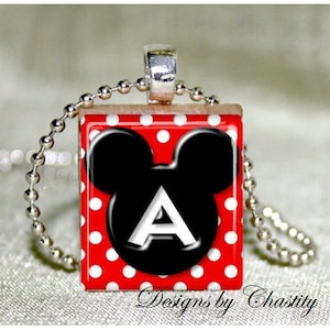 May include: A square pendant necklace featuring a red background with white polka dots. A black Mickey Mouse head shape with the white letter "A" is centered on the pendant. The necklace has a silver chain and the text "Designs by Chastity".