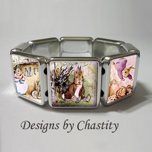 Op de afbeelding: Een zilverkleurige stretcharmband met vierkante panelen met illustraties van konijnen in een tuin. De armband heeft een gepolijste afwerking en een klassiek ontwerp. De tekst "Designs by Chastity" staat onderaan.