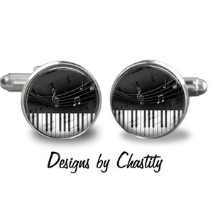 Può includere: Gemelli argentati con un design a tastiera di pianoforte in bianco e nero, sovrapposto a note musicali e una chiave di violino. Il testo "Designs by Chastity" è in basso.