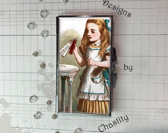 May include: A silver metal compact mirror featuring an illustration of Alice from Alice in Wonderland holding a bottle labeled "DRINK ME." The design includes the text "Designs by Chastity."