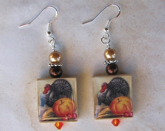 May include: A pair of dangle earrings featuring a Thanksgiving theme. Each earring has a square pendant with a turkey and pumpkin illustration, accented with brown and gold beads, and a red crystal charm.